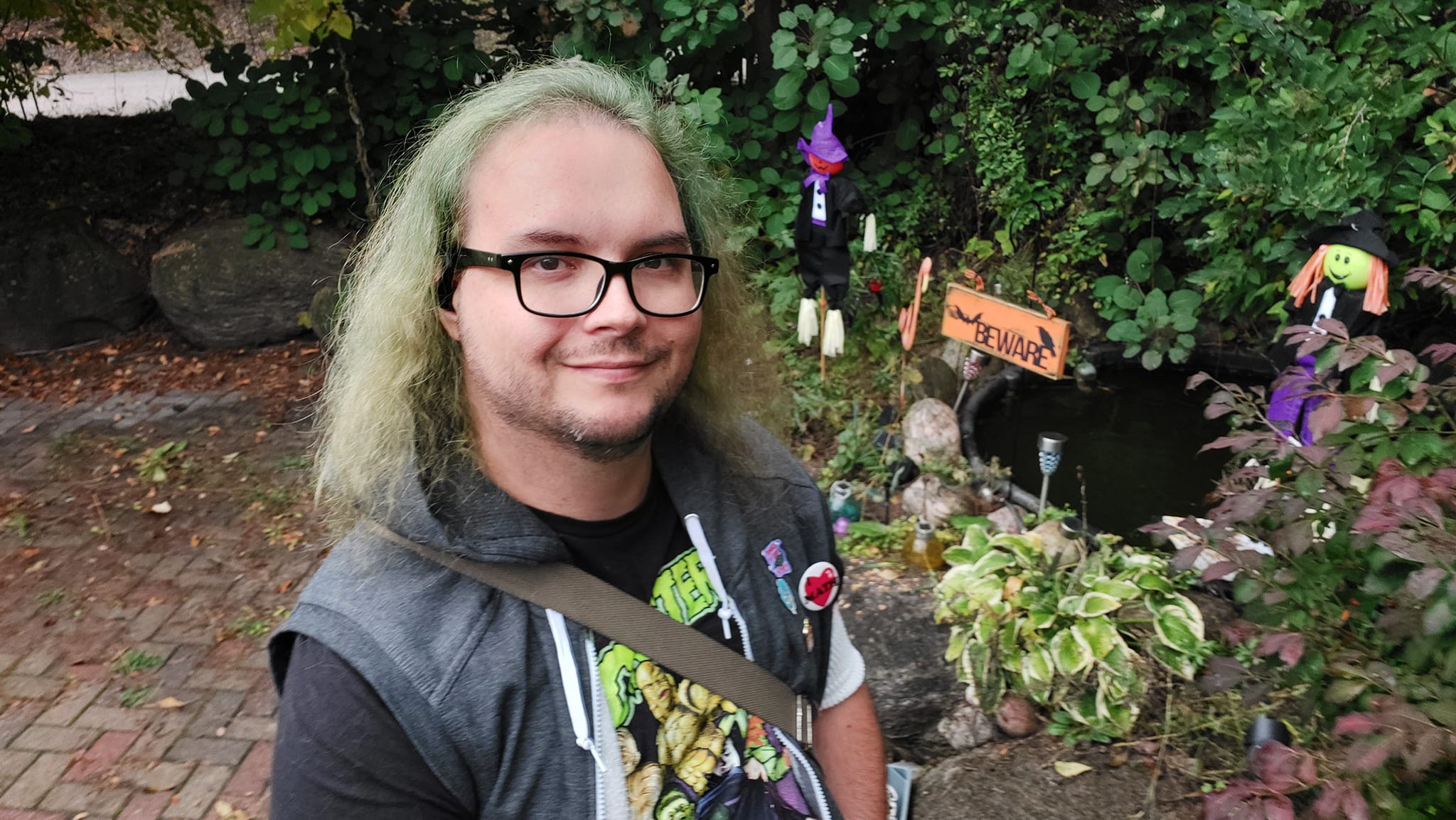 A man with long green hair stands on a patio, wearing an athletic sweater vest with some pins on it, a Universal Monsters shirt, and a messenger bag